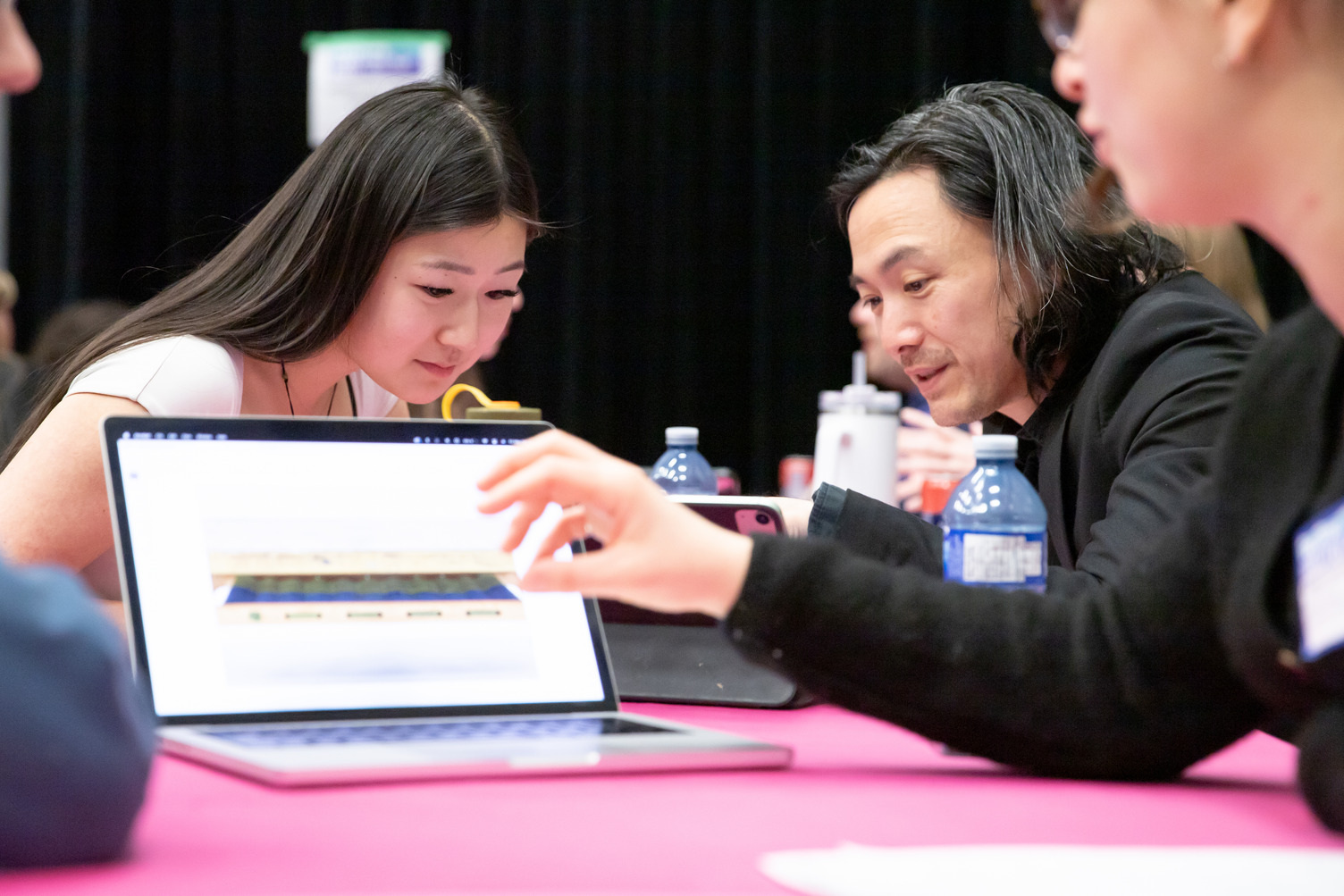 Two students and an industry lead sit at a table looking at a laptop