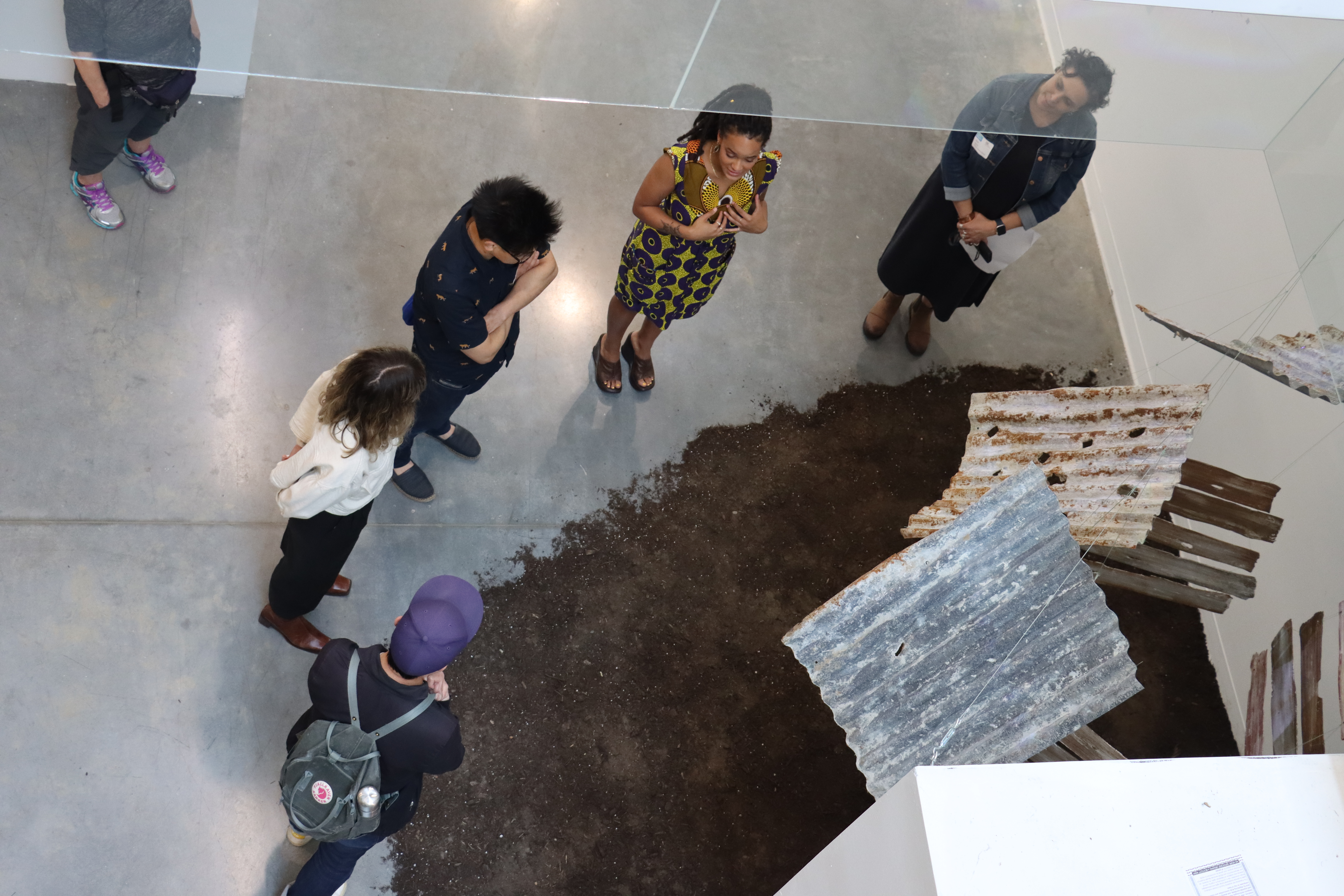 A shot from above of an artwork depicting grey roofing tiles with soil underneath - with five people surrounding it.