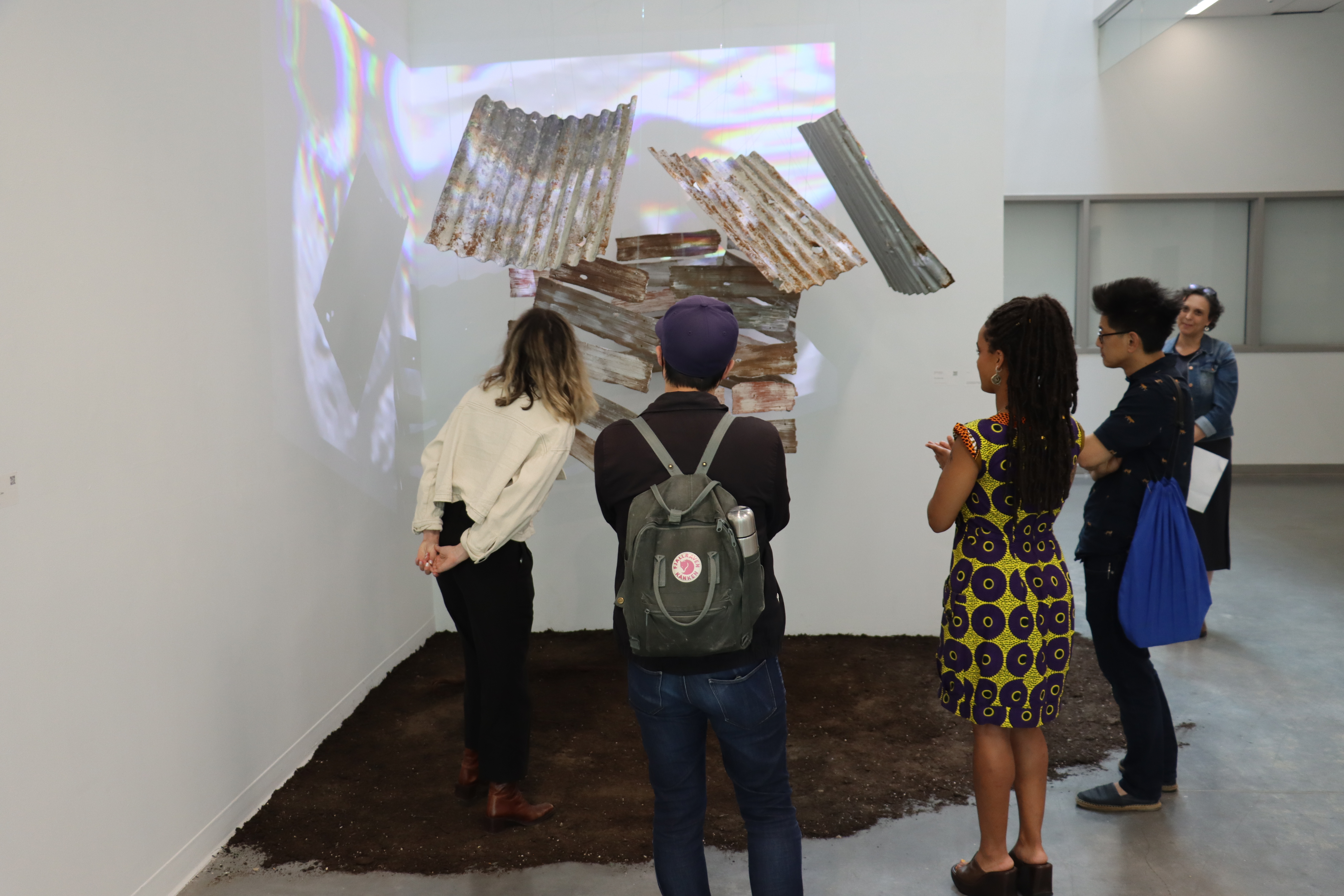Five people surrounding an artwork with grey metal roofing tiles and a video played on top of it