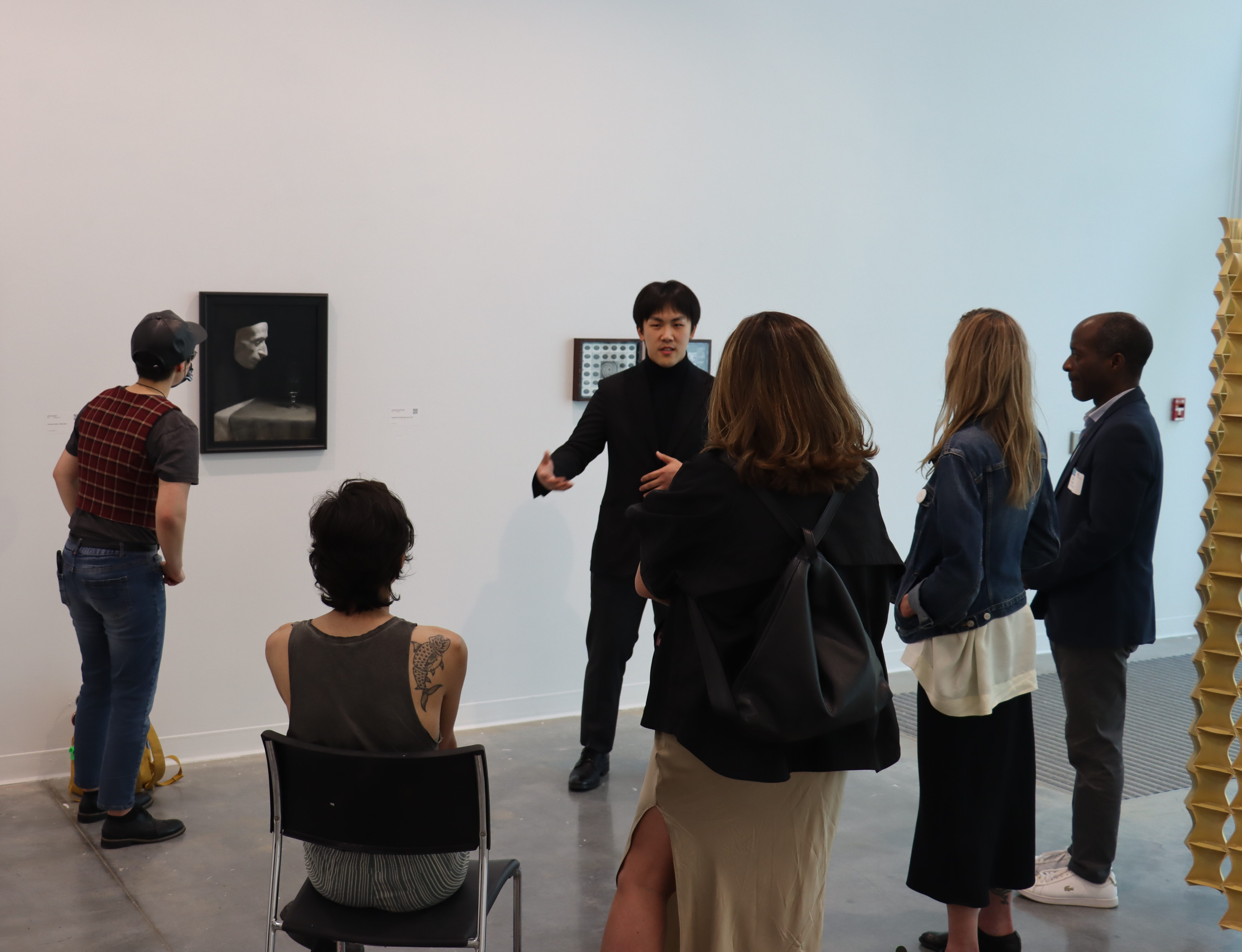 Four people surrounding an artist who is pointing to a painting of a floating mask above a grey tablecloth