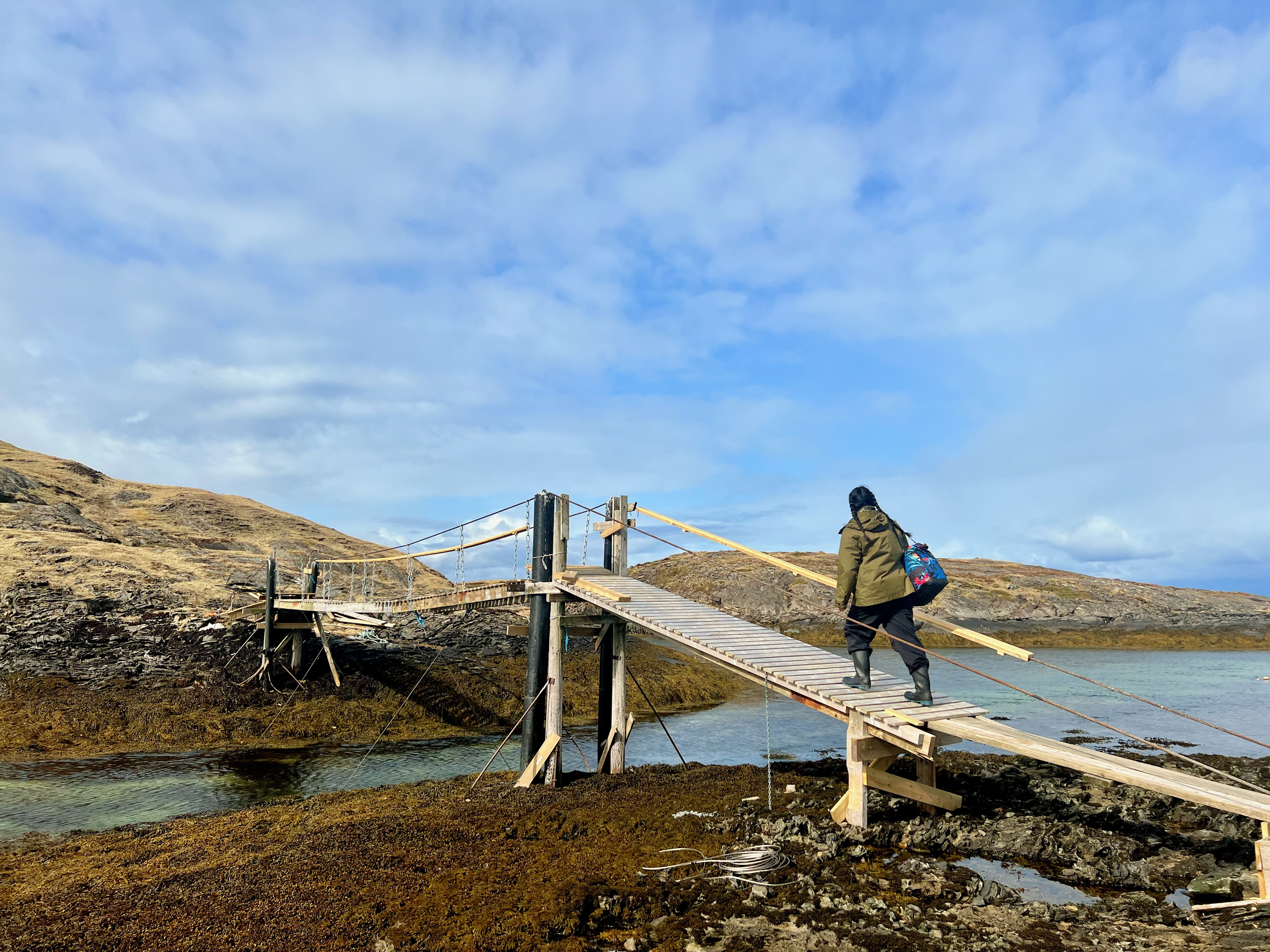 A person in the centre walking across a reedy slatted bridge among the hills