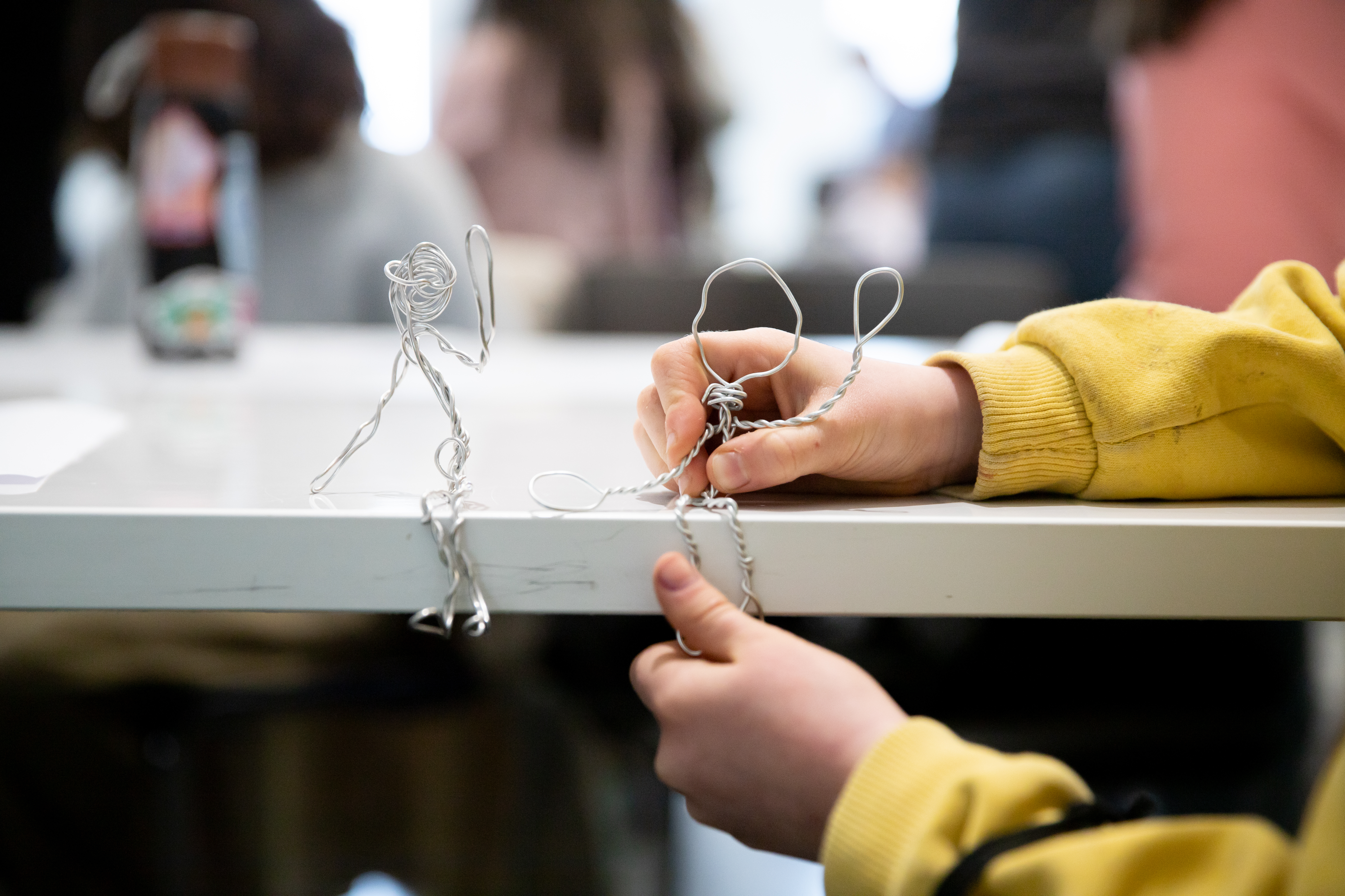 A students hand in frame with two wire stick figures sitting on a table
