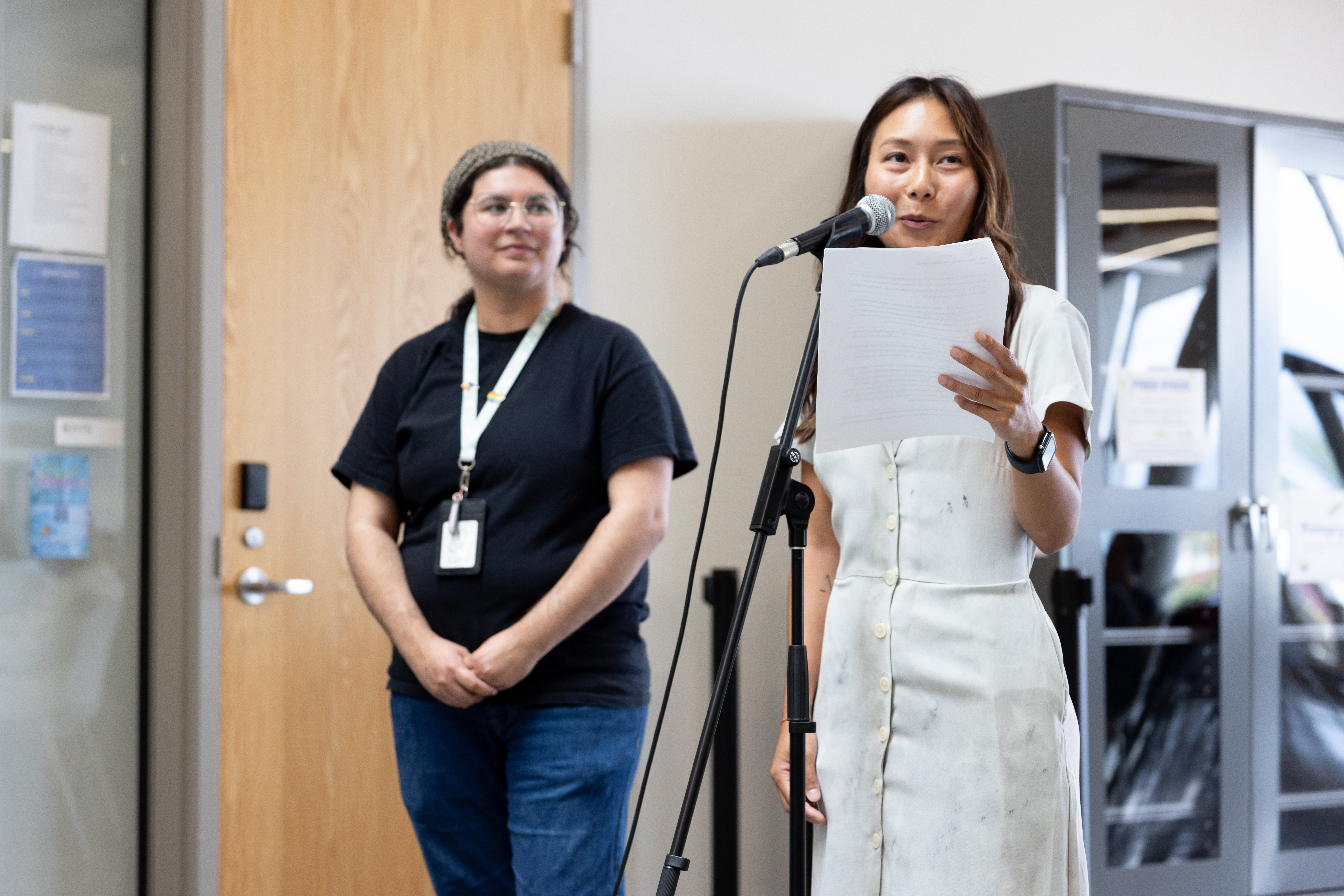 One person on the left looks on at a person holding a paper and talking at a microphone