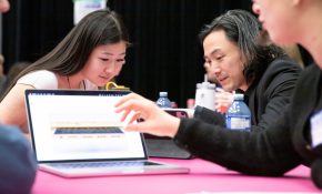 Two students and an industry lead sit at a table looking at a laptop