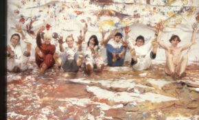 A group of students sit against a wall covered in painted handprints, playfully lifting up paint-covered hands and feet to the camera.