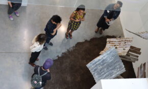 A shot from above of an artwork depicting grey roofing tiles with soil underneath - with five people surrounding it.