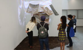 Five people surrounding an artwork with grey metal roofing tiles and a video played on top of it