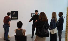 Four people surrounding an artist who is pointing to a painting of a floating mask above a grey tablecloth