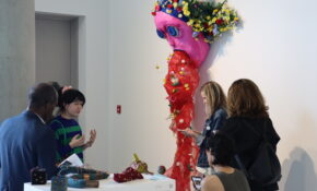 Four people surrounding a pink and red artwork that depicts a figure vomiting