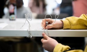 A students hand in frame with two wire stick figures sitting on a table