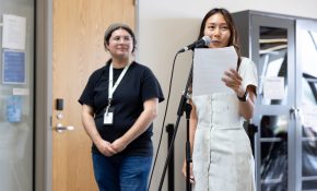 One person on the left looks on at a person holding a paper and talking at a microphone