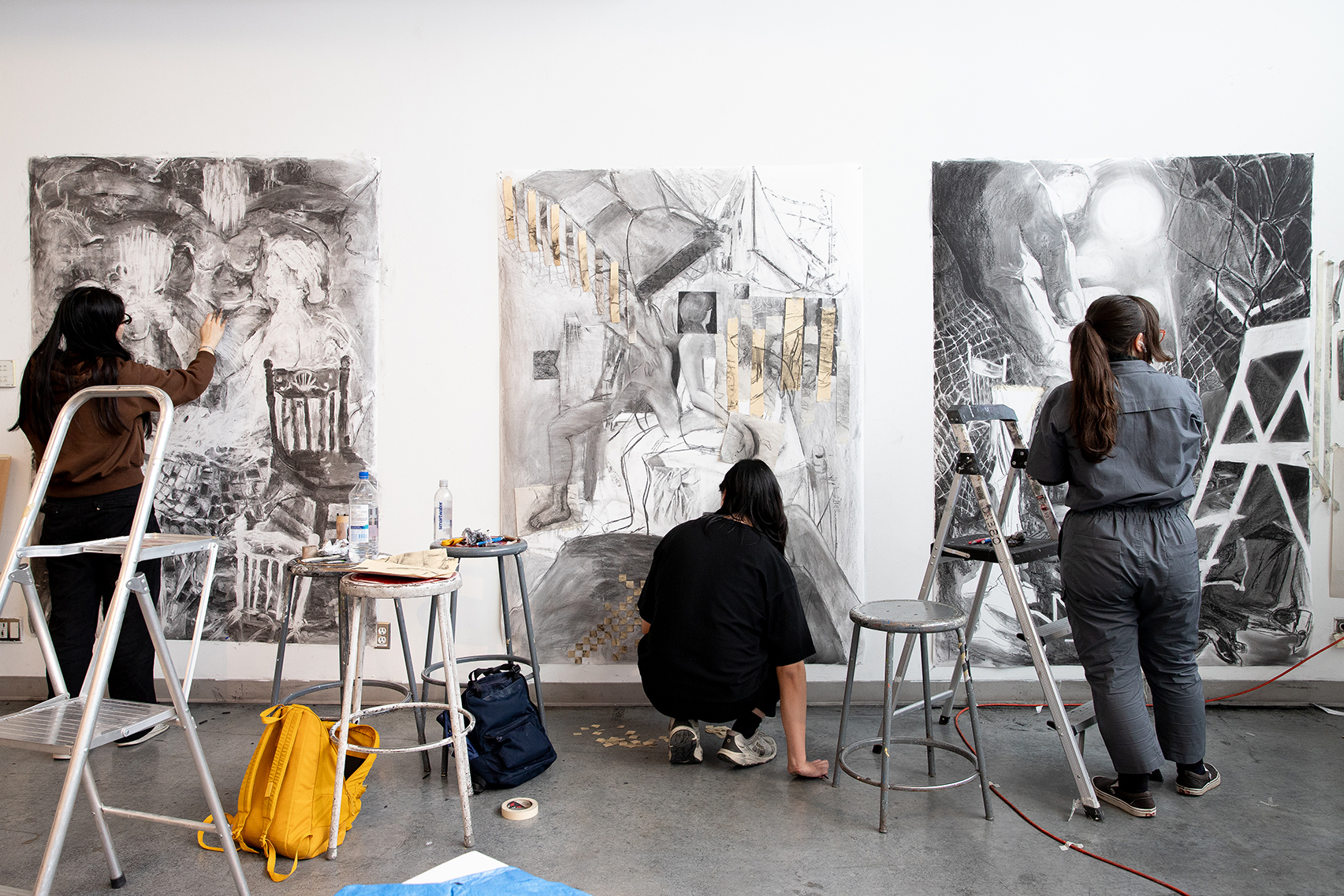 Three people work on large black-and-white drawings side-by-side in a studio.