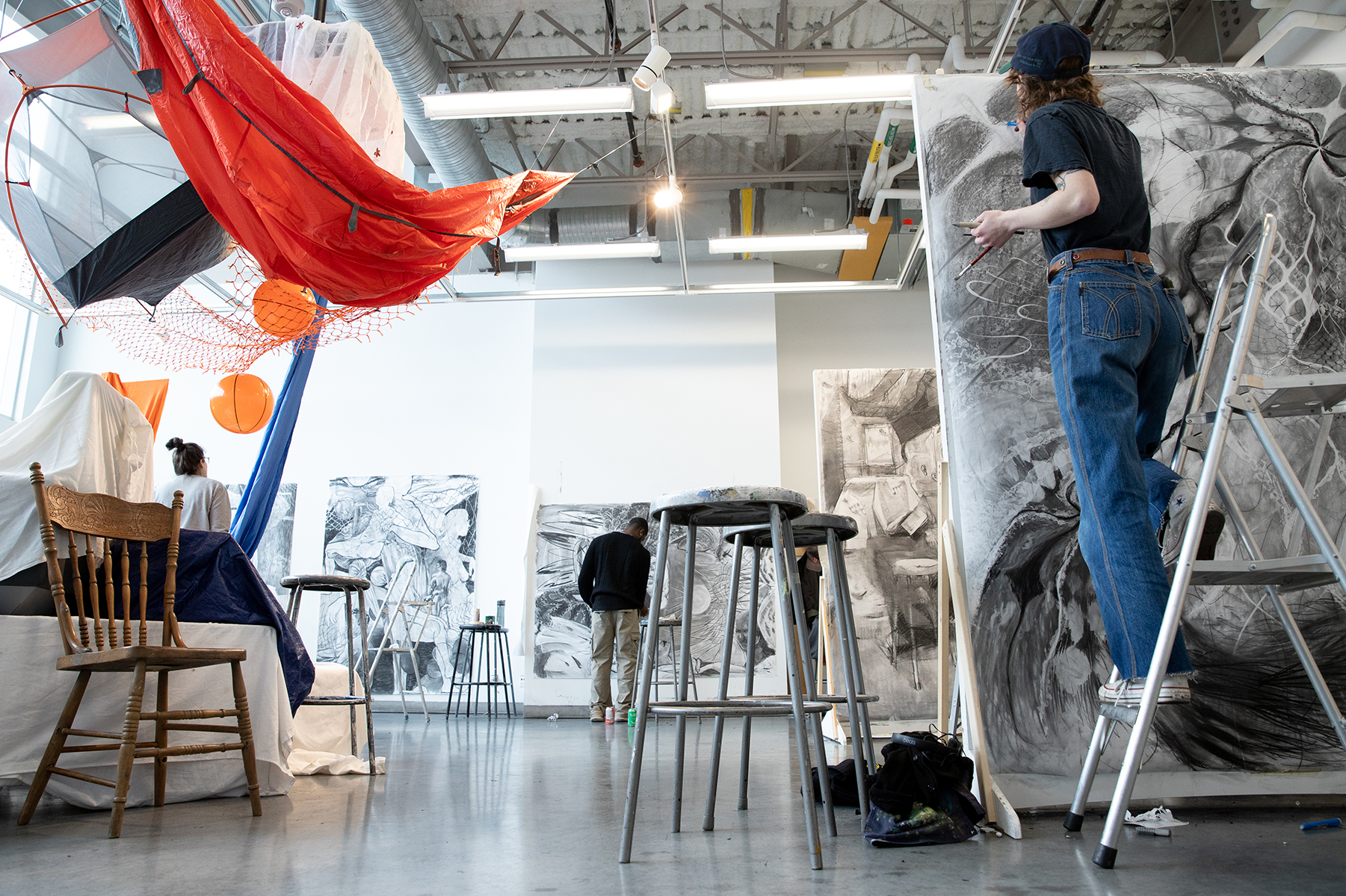 A still-life comprising furniture, netting and drapery hangs in a studio amidst a group of artists working on drawings.