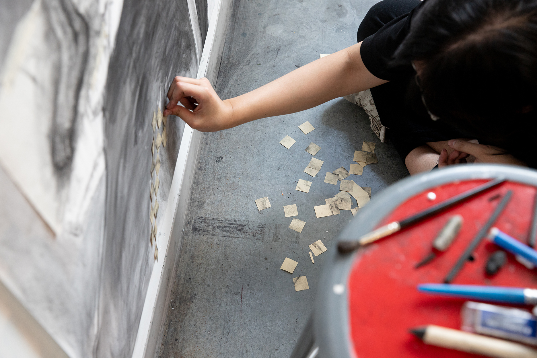 A person carefully applies small squares of collaged paper to a large black-and-white drawing.