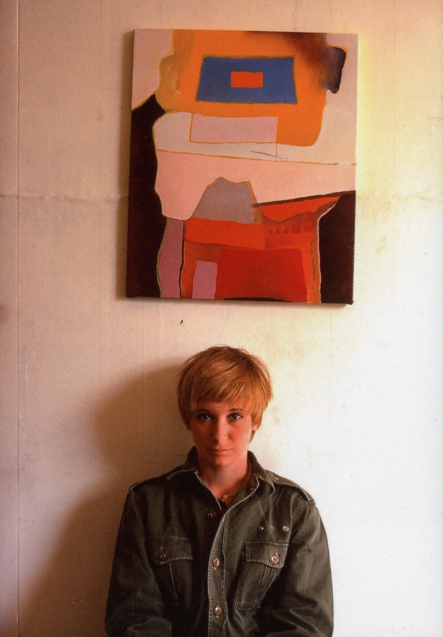 Young Janey Adams with short blonde hair, against a white wall with an abstract painting hanging overhead