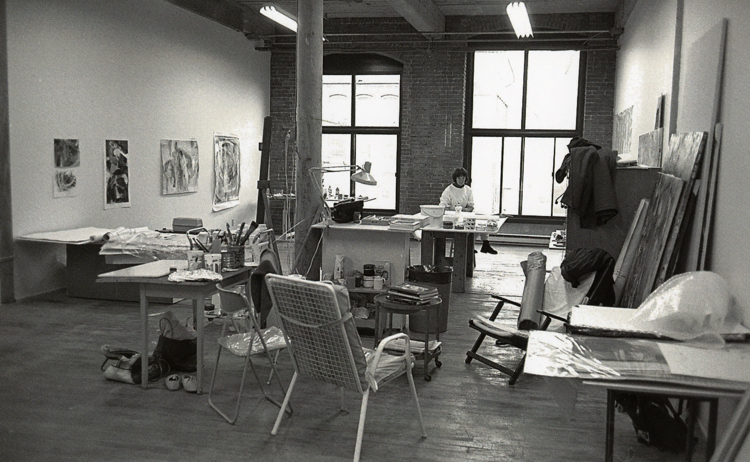 Black-and-white photo of an artist working in a spacious studio with brick walls and large windows.