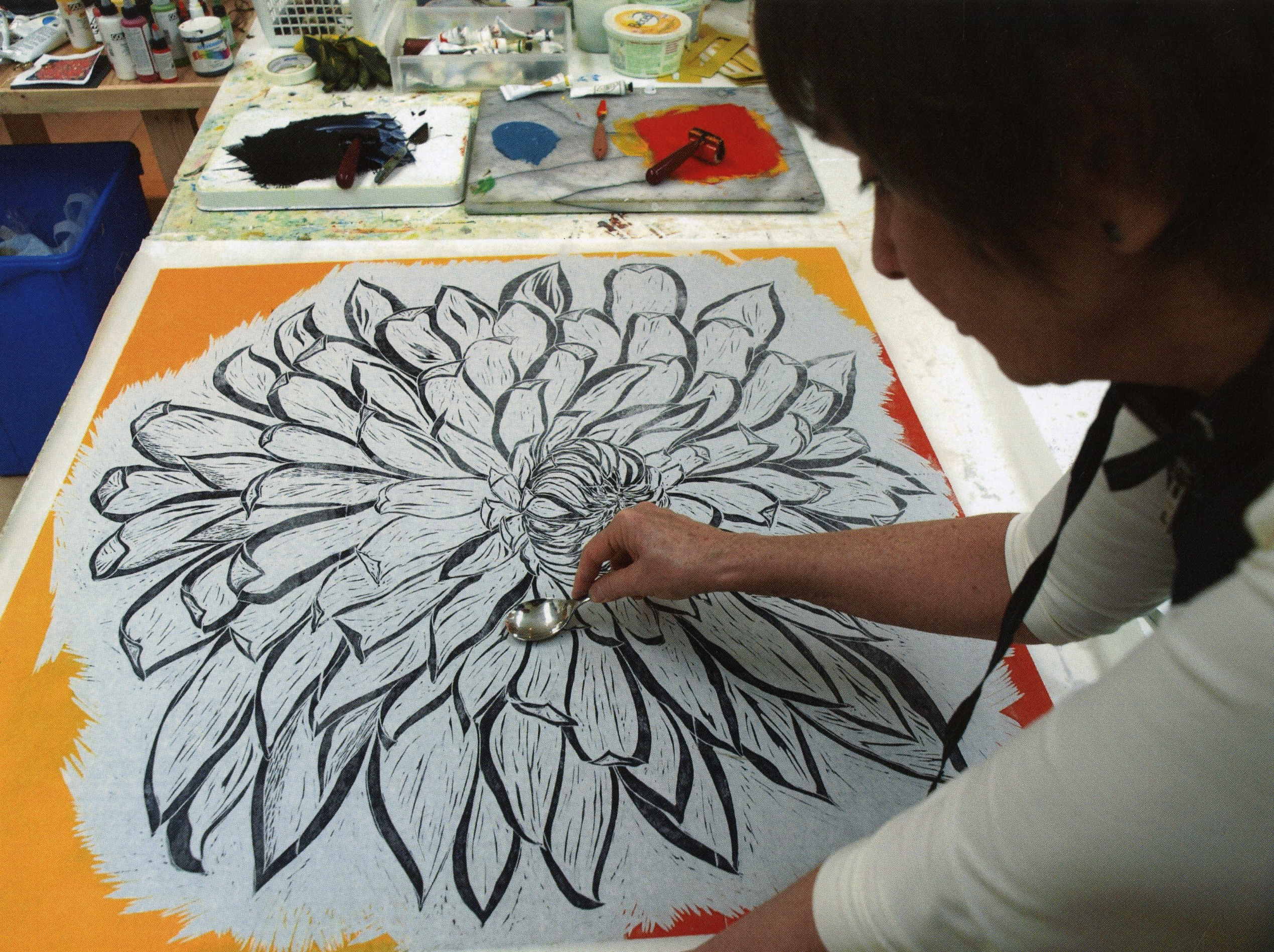 Artist using a spoon to burnish a large black-and-white flower print on a bright orange background.