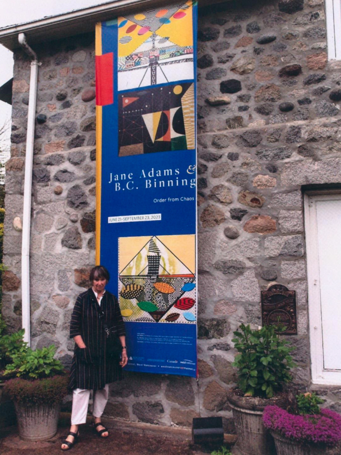 Person standing outside a stone building beside a tall blue exhibition banner reading “Jane Adams & B.C. Binning: Order from Chaos,” featuring colourful geometric artwork.