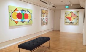 Bright gallery interior featuring geometric abstract paintings in vivid colours on white walls, with a dark bench centred on a polished wood floor.