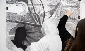 A person with long hair and a black shirt works on a black-and-white drawing.