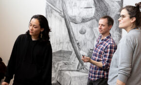Three people look together a piece of artwork.