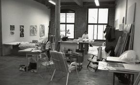 Black-and-white photo of an artist working in a spacious studio with brick walls and large windows.