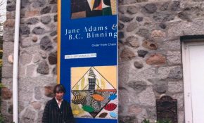 Person standing outside a stone building beside a tall blue exhibition banner reading “Jane Adams & B.C. Binning: Order from Chaos,” featuring colourful geometric artwork.